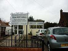 A flat-roofed temporary building,neatly finished in cream.  A ramp for 'disabled access' leads to the door, and two cars are parked on the right.  Prominent on the left a sign decorated with 'wrought' iron scrolls reads Bottesford town council, TEA POT HALL, pop-in room for the benefit of senior citizens