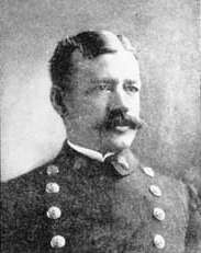 Head of a white man with neatly combed hair and a bushy mustache wearing a double-breasted military jacket with two columns of large buttons down the chest.