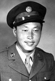 Head and shoulders of a smiling young man wearing a peaked cap and, over a shirt and tie, a military jacket with a round pin on each lapel.