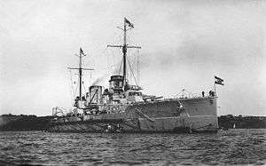 A large gray ship in port. The two funnels in the center of the ship emit clouds of smoke.