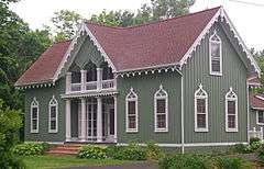 A green house with a reddish roof pointed on the front and the side. It has vertical raised wood on the sides and decorative flourishes above the windows and at the roofline.