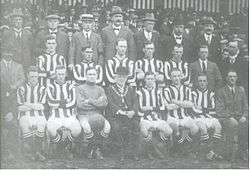 A football team comprising ten players in striped shirts and one in a shirt of a single colour pose for the camera. Five of the men are standing and the other six seated in front of them. Also posing with them are an elderly man in a bowler hat with a chain of office around his neck, and twelve men in business suits, some of whom are wearing hats. A crowd of spectators is visible behind the group.