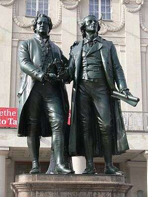 Photograph of a statue of Goethe and Schiller standing side by side, each looking forward. The statue is in front of the stone facade of an elaborate building. They are of nearly the same height. Goethe appears middle-aged; Schiller is noticeably younger. They are dressed in nineteenth century clothing. Goethe is wearing a knee-length formal coat, and Schiller is wearing a calf-length coat. Both men wear breeches. Goethe's left hand rests lightly on Schiller's shoulder; his right hand holds a laurel wreath near his waist. Schiller's right hand is nearly touching the wreath, which may suggest that Goethe is passing the wreath to Schiller. Schiller's left hand extends loosely below his waist, and grasps a rolled sheet of paper.