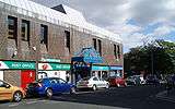 The side entrance of a 1970s brown shopping centre. A post office and charity shop can be seen.