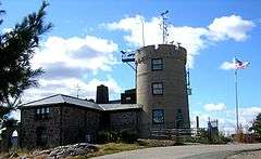 A two-story stone L-shaped building is attached to a round three-story tower with crenellated top. The tower has equipment mounted on the top, and antennae are visible in the background.