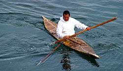 Man sitting with legs covered in boat that tapers to a point at each end holding long, pointed, wooden pole