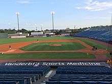 A view looking over the dirt and green grass of the infield as men rake the dirt and paint the lines before a game.