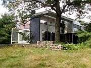 A strongly rectangular modern two story house. The exterior is brick, white tongue-in-groove vertical siding, and large glass windows. A tree and mortared stone wall stand in front of the house.