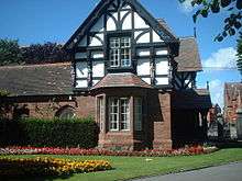 A two-storey house, the lower storey being in sandstone and the upper timber framed and gabled.  To the right of the house are the entry gates to the park and in front of it are formal flower beds.