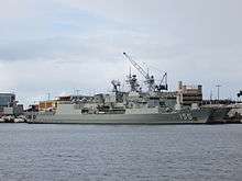 Colour photo of two grey-painted warships moored alongside a wharf. A large crane and several buildings are visible behind the ships.