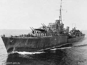 Photograph of a war ship in a body of water. Large cannons can be seen on the deck, along with sailors.