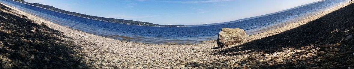 A boulder sits on a cobbled beach in bright sunlight with a stretch of blue water behind it. On the horizon are low forested hills. There is a snow covered volcano on the far distance. A few sailboats are in the water.