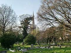 a number of grave stones in the grass, in the background trees with bare limbs and behind them a church with a tall thin spire