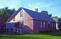 A  1 1⁄2-story wooden house, painted red, lit by the sun from the left with trees and part of a white house behind it.