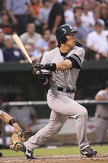 A man in a grey baseball uniform and blue batting helmet swings a baseball bat standing at home plate.