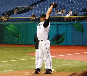 A Japanese man wearing a Devil Ray baseball uniform points his arms upward as he prepares to pitch in the bullpen.