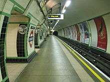 View along a platform in a circular tunnel. Tracks run on the right side with the walls covered with cream and green tiles. An illuminated "way out" sign overhead indicates the exit.