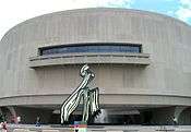 A large, convexly curving, 3 story tall, gray wall punctured on the second floor by a long strip of windows and a short balcony. A large black and white sculpture of a paint stroke sits in front of the building, stretching from the ground to the balcony