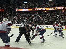 An official wearing black and white stripes conducts a faceoff as several players from two teams anticipate the start of play.