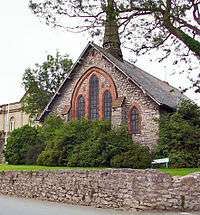 The end of a chapel with five windows and a small spire; in front is a bush and a wall