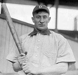 A man in a dark baseball cap and white shirt with a dark collar holds a baseball bat in both hands.