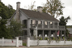 A light gray wooden house, five bays wide, with darker gray shutters, seen from its front right. It has a second-story balcony across the front supported by wooden pillars, two brick chimneys and a white picket fence with open gate in front.