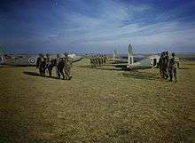 Three groups of six men wearing helmets and backpacks walk across a grass field towards waiting aircraft