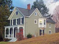 A beige-colored two-and-a-half-story wooden house with clapboard siding and black-and-white trim. Its roofs are pointed, with pointed-arch windows beneath them. In the middle of the center is a projecting oriel window. At left is an ornate porch.