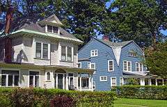 A white house and a blue house next to each other on a sunny morning with trees behind them