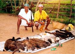 Two Malagasy hunters stand near a stream, one holding a gun, the other holding a lemur with a white head.