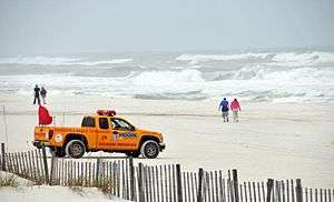 A Pensacola Beach Lifeguard pickup truck parked at a beach with rough seas. Two people are walking close to the water's edge in the background and two more are walking towards the water in the center of the image.