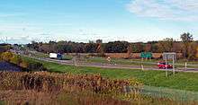 A divided highway going across a level landscape. On the right is a sign in English and French saying "Last U.S. Exit/Derniere Sortie EE. U." On the left the road goes away from the camera, up a slight rise to a more built-up area with a tall antenna.