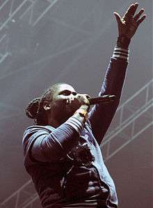 Man wearing dreadlocks in a bun singing into microphone, arm outstretched