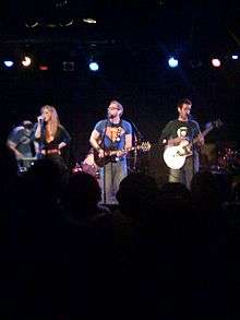 Photograph of a group singing and playing instruments on stage.