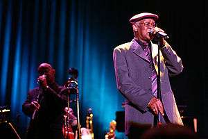 A man on the left wearing a black suit and holding a standup microphone and another man with glssses on the right with a grey suit and a red benet holding a microphone.