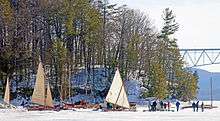 Several boats with their sails at various stages of dismantling surrounding a large vehicle with a wooded promontory behind them. In the far right upper background a portion of a bridge is visible