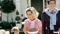  Image of a blond woman. She is a housewife and carries a pie in her left hand. Her hair is styled in 1950s fashion. She is wearing a pink polo shirt. Surrounding her are her husband and children.