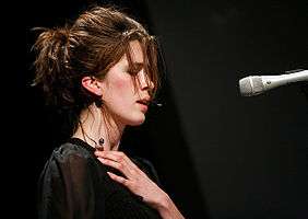 Imogen Heap face-on, on 12 October 2006 at the Carling Academy Birmingham, at a multi-tier set-up with keyboard atop which she's intently playing.  She has a headset microphone, then-characteristically back-combed hair, wears a sleeveless frilly pink bodice and is rear illuminated by two piercing green star-burst spotlights.