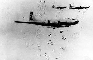 Black and white photo of three multi-engined aircraft flying in formation while dropping a large number of bombs