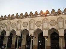 Five keel shaped arches (and part of a sixth) are visible. The arches are supported by cylindrical columns. Above each arch is a large circular inscribed stucco decoration, and above each column is a large inscribed stucco decoration that mirrors the shape of arch and columns. Behind the row of columns is a walkway, and then a wall with entrances the shape and size of the arches and columns.