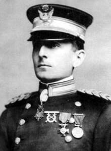 Head and shoulders of a white man standing erect, wearing a peaked cap over his eyes and a double-breasted military jacket with thick braided cords on the shoulders, three medals on the left breast, and a star-shaped medal hanging from his neck.