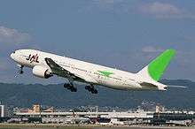 A Boeing 777–200 aircraft in mid air during take-off, with the view of Itami Airport in the background