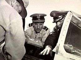 Three men in military uniforms with peaked caps crowding around the open cockpit of a military aircraft, two facing the camera and one with his back turned. The central figure in the portrait, facing the camera, has two rows of braid on the visor of his cap, and pilot's wings on the left breast pocket.