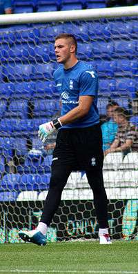 A young white man with close-cropped dark hair, wearing a blue sports shirt, black shorts and leggings, and white gloves, walks in front of a football goal.