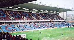 A two-tiered cantilever football stand. The lower tier has light blue seats with some claret seats which spell the word "Clarets". The upper has all claret seating. Some floodlights are attached to the roof of the stand. A scattering of spectators can be seen in the seats.