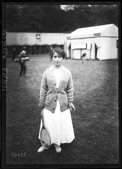 A woman standing straight on at the camera angle with a colored sweater on with all white attire and a tennis racket in right hand, which this picture is a black and white