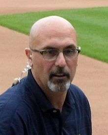 A close-up of a man wearing glasses and a navy blue polo shirt on a baseball field