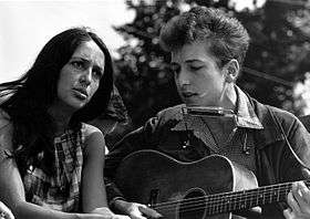 medium shot of a woman with long dark hair on left and man playing an acoustic guitar on the right