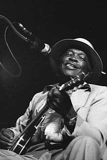 A black-and-white photograph of a man performing electric guitar in a grey suit and white hat; he is seated. A microphone is visible in front of him.