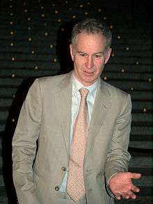 A grey-haired man in a grey suit and tie raises his left hand